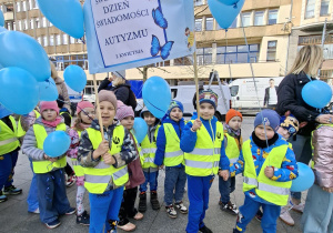Dzieci stoją na Pasażu Schillera w Łodzi ubrane w żółte kamizelki trzymają niebieskie balony z helem i transparenty które głoszą hasła zrozumienia dla dzieci w spektrum autyzmu.
