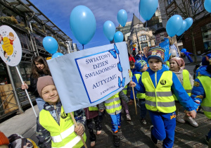 Dzieci stoją na Pasażu Schillera w Łodzi ubrane w żółte kamizelki trzymają niebieskie balony z helem i transparenty które głoszą hasła zrozumienia dla dzieci w spektrum autyzmu.