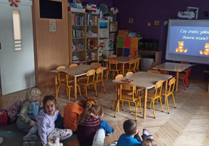 Dzieci siedzą na dywanie i oglądają bajkę o misiu na tablicy interaktywnej