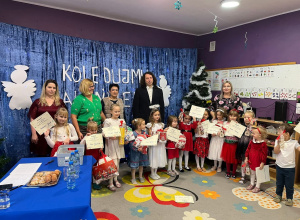 Dzieci wraz z jury konkursu Kolędujmy Maleńkiemu pozują do zdjęcia grupowego na tle dekoracji świątecznej