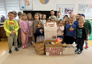 Przedszkolaki trzymają plakat PCK dotyczący zbiórki, przed dziećmi są zgromadzone dary, które zebrały dzieci wraz z rodzicami.