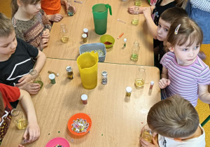 dzieci siedzą przy połączonych stolikach, na stołach są koraliki, brokat i inne atrybuty do zrobienia butelek sensorycznych.