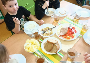 Dzieci siedzą przy zastawionych stolikach. Na stole widnieje pełnowartościowe pieczywo, warzywa i owoce, owsianka