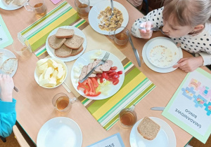 Dzieci siedzą przy zastawionych stolikach. Na stole widnieje pełnowartościowe pieczywo, warzywa i owoce, musli śniadaniowe. Dzieci również trzymają kartki z napisem Śniadaniowe bingo