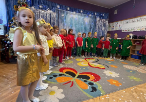 dzieci ubrane w zielone i czerwone stroje stoją w półkolu podczas przedstawienia świątecznego dla rodziców