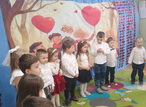 Dzieci w białych koszulach i ciemnych spodenkach i spódniczkach stoją na tle dekoracji na Dzień Babci i Dziadka, śpiewają piosenkę.