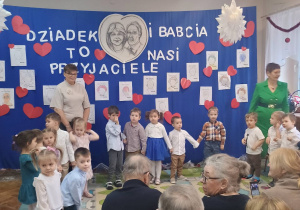 Dzieci z najmłodszej grupy stoją w półkolu wraz ze swoimi nauczycielkami na tle dekoracji z okazji Dnia Babci i dziadka