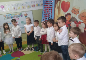 Dzieci w białych koszulach i ciemnych spodenkach i spódniczkach stoją na tle dekoracji na Dzień Babci i Dziadka, śpiewają piosenkę.