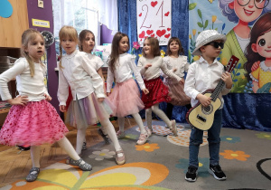Grupka dziewczynek w różowych spódniczkach stoi w tanecznej pozie, chłopiec w ciemnych okularach udaje, że gra na gitarze
