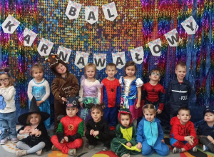 zdjęcie grupowe dzieci z grupy Maluszków na tle napisu "Bal Karnawałowy" i kolorowym materiale.