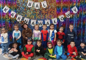 zdjęcie grupowe dzieci z grupy Maluszków na tle napisu "Bal Karnawałowy" i kolorowym materiale.