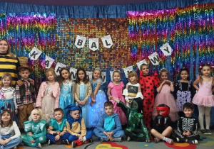 zdjęcie grupowe dzieci z grupy Starszaków na tle napisu "Bal Karnawałowy" i kolorowym materiale.