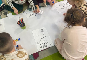 Dzieci z grupy Maluszków kolorują wspólną kolorowankę dinozaurową, prace wykonują na dywanie kredkami