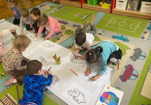 Dzieci z grupy Maluszków kolorują wspólną kolorowankę dinozaurową, prace wykonują na dywanie kredkami