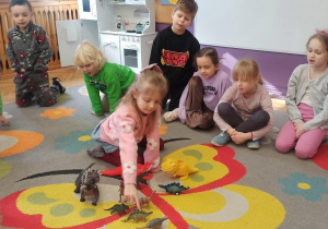 Dzieci siedzą w kręgu, jedna dziewczynka jest w środku koła i układa dinozaurowi figurki