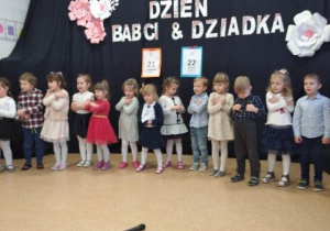 Grupa dzieci w odświętnych strojach na tle dekoracji z napisem "Dzień Babci &Dziadka" w trakcie występu.