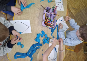 Dzieci siedzą przy stolikach i wykonują pracę plastyczną, wyklejają niebieską bibułą kropelki wycięte z papieru