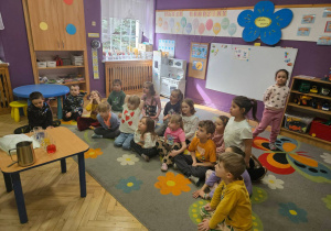 Dzieci siedzą na dywanie i obserwują jakie procesy zachodzą podczas eksperymentu z wodą wykonywanego przez nauczyciela