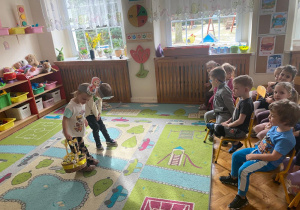 Dwoje dzieci stoją na dywanie i kłaniają się, reszta dzieci siedzi na krzesłach jako widownia