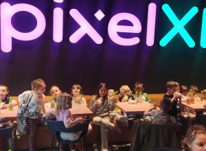 dzieci siedzą przy stolikach, na stolikach widać butelki z sokiem i drugie śniadanie. Nad dziećmi jest neonowy napis PIXEL XL