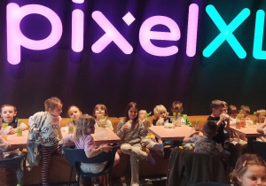 dzieci siedzą przy stolikach, na stolikach widać butelki z sokiem i drugie śniadanie. Nad dziećmi jest neonowy napis PIXEL XL
