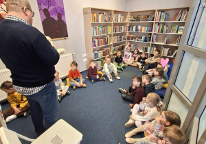Dzieci siedzą na dywanie w przestrzeni edukacyjnej biblioteki i słuchają pana bibliotekarza
