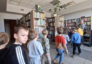 Dzieci w kolejce idą z panem bibliotekarzem i zwiedzają bibliotekę.