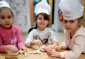 Trzy dziewczynki siedzą przy stoliku w jadalni. Pozują do zdjęcia przygotowując składniki do pizzy. Na głowach mają kucharskie czapki.