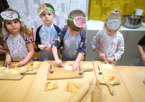 Czworo dzieci w fartuszkach i opaskach z pizzą na głowie stoją przy stoliku i kroją składniki na pizzę.