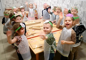 Zdjęcie grupowe. Dzieci stoją dookoła stołu. Ubrane są w fartuszki i opaski na głowę z obrazkiem pizzy. Na stole leżą przybory - wałki do ciasta i deski do krojenia.