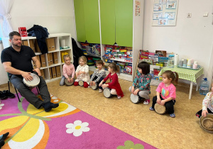 Dzieci siedzą na bębnach i uczą się grać. Z boku zdjęcia siedzi pan prowadzący warsztaty i grający na bębnie.