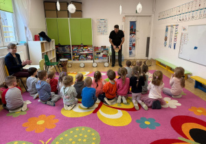 Dzieci siedzą tyłem na dywanie a przed nimi stoi pan, który prezentuje sposób gry na bębnach afrykańskich