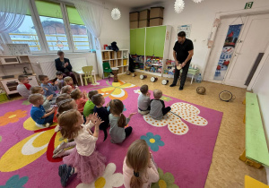 Dzieci siedzą tyłem na dywanie a przed nimi stoi pan, który prezentuje sposób gry na bębnach afrykańskich