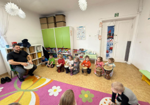 Dzieci siedzą na bębnach i uczą się grać. Z boku zdjęcia siedzi pan prowadzący warsztaty i grający na bębnie.