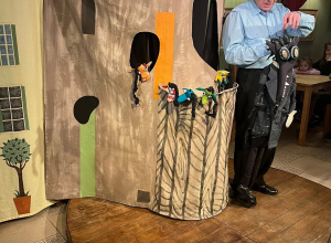 Na zdjęciu przedstawiono scenę teatralną. Mężczyzna trzyma dużą kukłę przypominającą smoka, a w tle dzieci oglądają przedstawienie. Widoczne są dekoracje przypominające zamek.