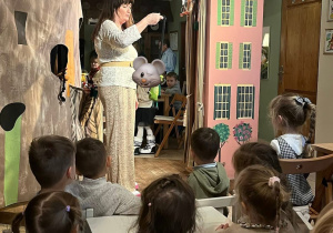 Na zdjęciu widać dzieci siedzące przy stolikach i oglądające przedstawienie teatralne w Baśniowej Kawiarence. Przed dziećmi stoi kobieta prezentująca lalkę teatralną. W tle znajdują się kolorowe dekoracje przypominające kamienice oraz scena teatralna. Atmosfera jest ciepła i przyjazna.