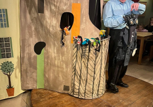 Na zdjęciu przedstawiono scenę teatralną. Mężczyzna trzyma dużą kukłę przypominającą smoka, a w tle dzieci oglądają przedstawienie. Widoczne są dekoracje przypominające zamek.