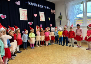 Dzieci stoją w półkolu przebrane za skrzaty.W tle jest dekoracja z okazji Dnia Babci i Dziadka.