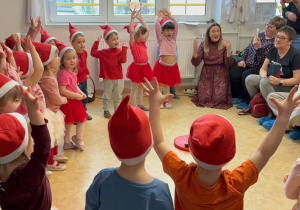 Dzieci stoją w półkolu przebrane za skrzaty.W tle jest dekoracja z okazji Dnia Babci i Dziadka.