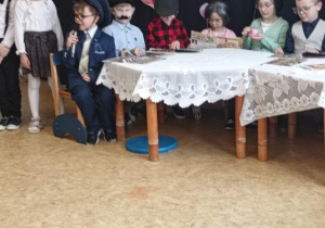 Dzieci z grupy straszaków występują podczas przedstawienia z okazji Dnia Babci i Dziadka. Dzieci są przebrane za babcie i dziadków, na scenie stoi stół przykryty białym obrusem, dzieci siedzą za stołem, w tle jest dekoracja z okazji Dnia Babci i Dziadka.