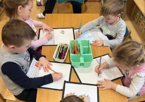 Dzieci siedzą przy stoliku i kolorują komputery, które tworzą z papieru.