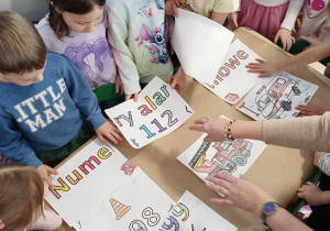 Dzieci siedzą przy stolikach i wspólnie przyklejają elementy pokolorowane przez siebie na duży arkusz papieru.