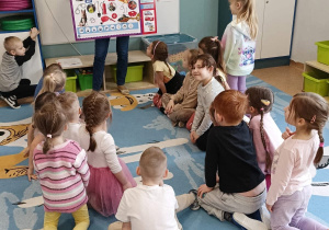 Dzieci siedzą na dywanie w kole, nauczyciel prezentuje tablice edukacyjną dotyczącą numerów alarmowych