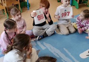 Dzieci siedzą w kole i trzymają w rękach kartki z numerami alarmowymi zapisanymi na czerwono.