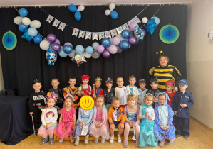 Dzieci z grupy średniaków pozują do zdjęcia grupowego wraz z nauczycielem. Każdy jest przebrany w strój karnawałowy, w tle widać dekoracje z okazji Balu.