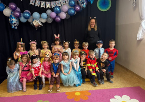 Dzieci z grupy maluszków pozują do zdjęcia grupowego wraz z nauczycielem. Każdy jest przebrany w strój karnawałowy, w tle widać dekoracje z okazji Balu.