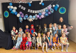Dzieci z grupy starszaków pozują do zdjęcia grupowego wraz z nauczycielem. Każdy jest przebrany w strój karnawałowy, w tle widać dekoracje z okazji Balu.