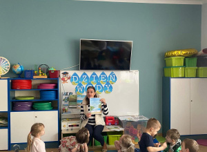 Nauczyciel prezentuje dzieciom kartkę edukacyjną dotyczącą wody. Dzieci siedzą w kole na dywanie i uważnie słuchają nauczyciela.