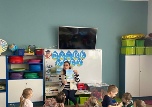 Nauczyciel prezentuje dzieciom kartkę edukacyjną dotyczącą wody. Dzieci siedzą w kole na dywanie i uważnie słuchają nauczyciela.