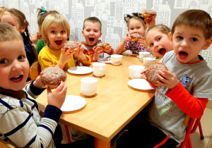 Zdjęcie grupowe. Dzieci z najmłodszej grupy siedzą przy stoliku w jadalni i jedzą pączki.